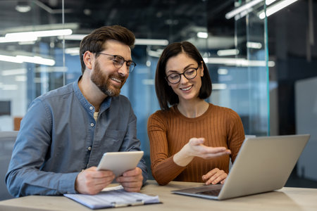 Two colleagues engaging in discussion, utilizing laptop and tablet for project collaboration in modern office environment. Man holding tablet, woman gesturing towards laptop screenの写真素材