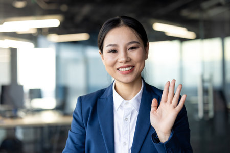 Confident woman wearing blue suit smiling and waving at camera during video call. Engages in professional communication in office environment. Concept of modern business, technology, connectionの写真素材
