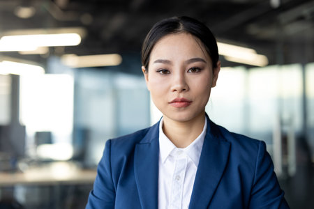 Confident businesswoman wearing blue suit looking directly at camera in modern office setting. Professional demeanor and sharp attire signify leadership and assertiveness in corporate environment.の写真素材