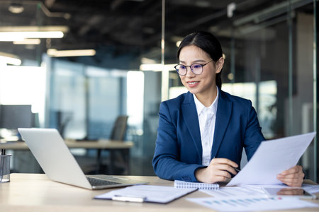 Focused Asian businesswoman working at office desk with laptop and documents. She analyzes papers, demonstrates professional business environment. Perfect for business, technologyの写真素材
