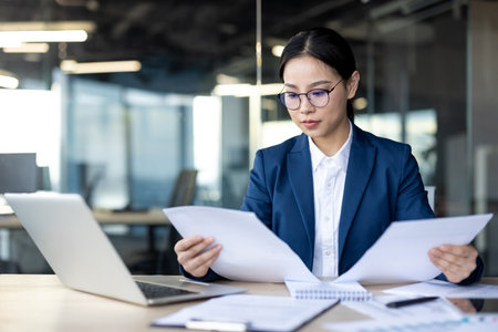 Focused financier reviews paperwork in office setting. Dressed in formal attire, analyzing documents at desk with laptop open nearby. Represents business professionalism and attention to detail.の写真素材