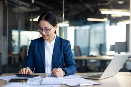 Asian businesswoman focuses on calculations using documents and laptop in office. She appears engaged and confident, reflecting professionalism and dedication.の写真素材