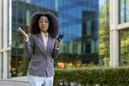 Hispanic businesswoman standing outside modern office building, showing confused expression and holding smartphone. Professional attire and natural surroundings emphasize urban business lifestyle.の写真素材