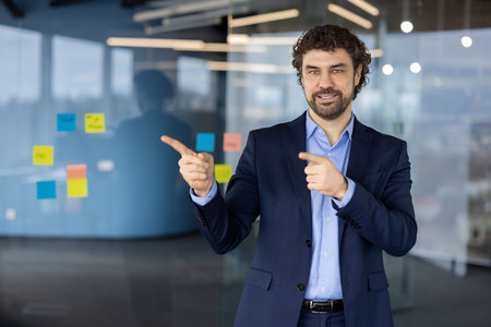 Businessman in office setting pointing at glass wall with sticky notes. Professional wearing blue suit projects confidence and positivity. Displays concept of teamworkの写真素材