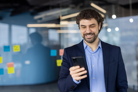Businessman wearing suit using smartphone in modern office. Man smiling, engaging with phone for work communication or tasks. Office environment with note-covered glass board in background.の写真素材