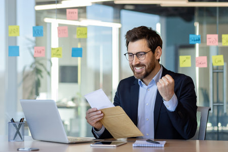 Businessman in suit happily reads successful notification from envelope at desk. Celebrates good news in contemporary office with notes and laptop. Concept of achievement, success, and communication.の写真素材