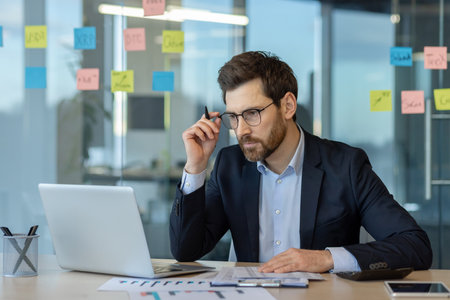 Businessman in suit intensely focuses on laptop for data analysis in modern office. Sticky notes on glass wall indicate brainstorming. Displays deep concentration and professional work environment.の写真素材