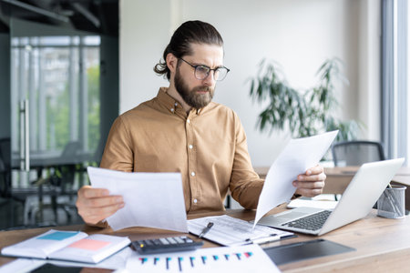 Confident male employee analyzing paperwork in contemporary office setting. Professional businessman using laptop for efficient work process. Serious focus on details and strategic planningの写真素材