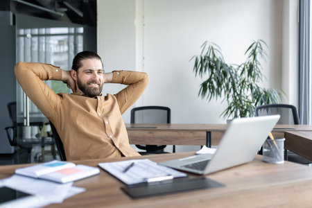 Confident businessman seated at desk with hands behind head, smiling while taking break. Modern office environment with natural light, laptop, and documents reflecting comfortable workplace.の写真素材