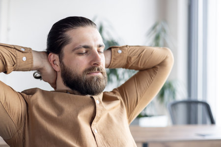 Bearded male office worker relaxing in contemporary office setting. Person enjoying a serene moment leaning back in chair, hands behind head, with closed eyes.の写真素材