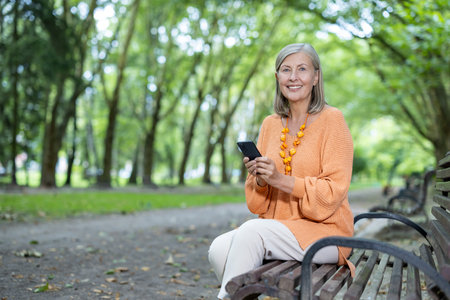 Mature woman smiling as she sits on bench in lush park setting, using her smartphone. Captures...