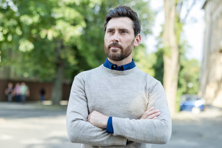 Confident adult male professional standing with arms crossed on urban street. Mature businessman exudes leadership and focus, dressed in casual attire. Outdoors setting highlights city environmentの写真素材