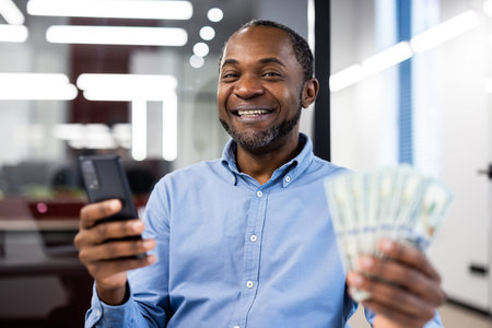 Smiling businessman holding stack of cash while using smartphone in office, showing financial...