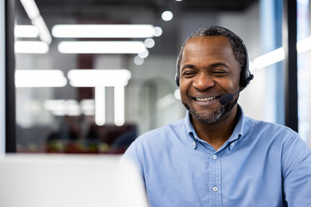 Confident African American mature businessman in virtual meeting. Smiling wearing headset, working in modern office environment. Focus on professional interaction, communication, and technology.の写真素材