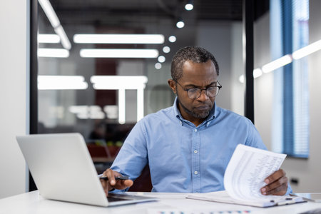 Business professional concentrating on reviewing documents at desk with laptop in modern office. Man...