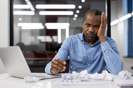 Mature African American businessman sitting at office desk looking stressed and overwhelmed. Holding...
