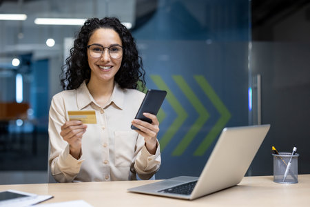 Smiling woman holding credit card and smartphone while sitting at desk with laptop. She wears...