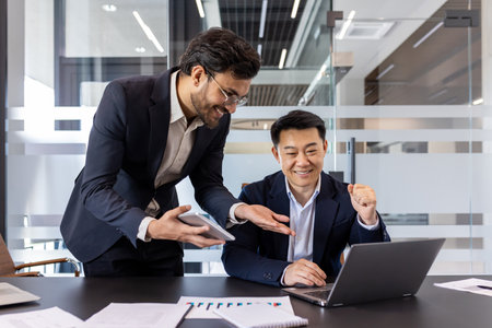 Business professionals in collaborative environment discussing ideas over laptop and smartphone. Men in formal attire engage in productive teamwork, showcasing excitement and achievement in officeの写真素材