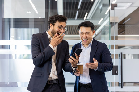 Two colleagues sharing a light moment in office while watching a funny video on smartphone. Both men dressed in business suits, one holding a coffee cup, expressing laughter and surprise.の写真素材