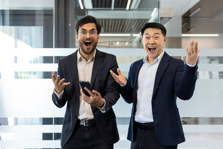 Two businessmen in formal attire expressing excitement and surprise in modern office environment. One holds smartphone, emphasizing technology and achievement. Perfect representation of teamworkの写真素材