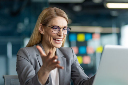 Confident professional woman expressing enthusiasm during virtual meeting on laptop. Wearing eyeglasses and formal attire, she communicates confidently, embodying teamwork and leadershipの写真素材