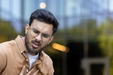 Young male clutching chest in discomfort indicating possible heart-related issue outdoors. Expression of pain and concern suggests sudden health condition.の写真素材