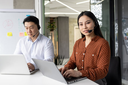 Asian business professionals using laptops and headsets in modern office environment. Engaged in teamwork, communication, and technology solutions.の写真素材