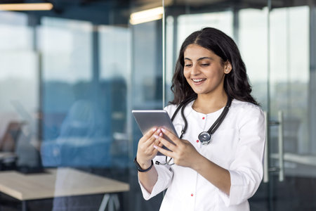 Smiling female doctor stands in modern office using digital tablet. Stethoscope around neck suggests...