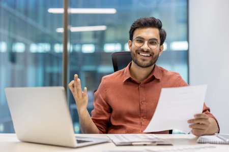 Confident businessman smiling while holding document at office desk. Displays positive energy and professionalism. Man engages with paperwork beside laptop, demonstrating workplaceの写真素材