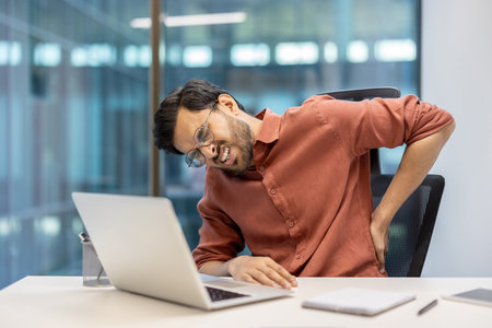 Man sitting at desk using laptop experiencing back pain. Wearing glasses shirt. Scene depicts discomfort from prolonged sitting. Represents office work, health issues, and need ergonomic solutions.の写真素材