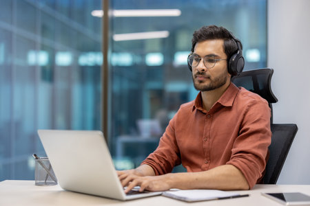 Young professional wearing headphones working on laptop in modern office environment. Casual attire and focused expression highlight dedication and productivity. Ideal for concepts of remote workの写真素材