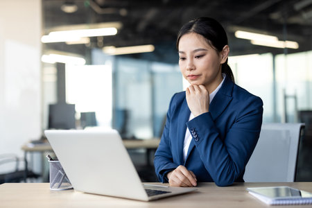Confident asian businesswoman wearing blue suit using laptop at office desk. Woman focused on work, reflecting professionalism, concentration. Modern office setting enhances productive atmosphere.の写真素材