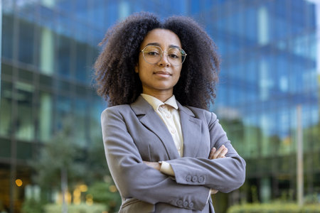 Confident African American businesswoman standing outside modern glass office building. Her expression conveys determination and leadership. Ideal for business, success, and empowerment concepts.の写真素材