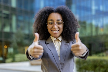 Confident African American business woman giving positive gesture outside office building. Professional attire, bright smile, symbolizing success, approval, motivation.の写真素材