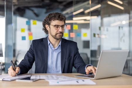 Mature financier sits at desk engaged in paperwork and laptop use. Focused on financial analysis and investment strategy in office environment. Professional emphasizes business planningの写真素材