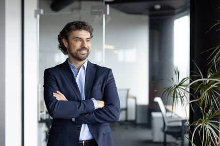 Confident businessman standing in modern office with arms crossed. He appears optimistic and focused, representing success, leadership, and future growth within corporate atmosphere.の写真素材