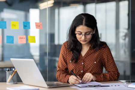 Young professional woman in office concentrating on paperwork, surrounded by laptop, and modern office setting. Scene conveys concentration, work, and productivity in professional environment.の写真素材