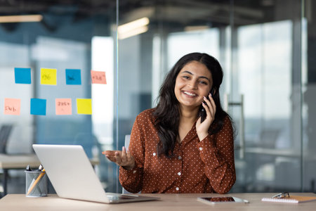Woman smiling talking phone in modern office. Laptop on desk with colorful sticky notes on glass...
