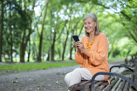 Senior woman seated on park bench using smartphone, smiling and enjoying outdoor leisure. Wearing casual , showing happiness and connectivity in nature setting with lush greenery.の写真素材