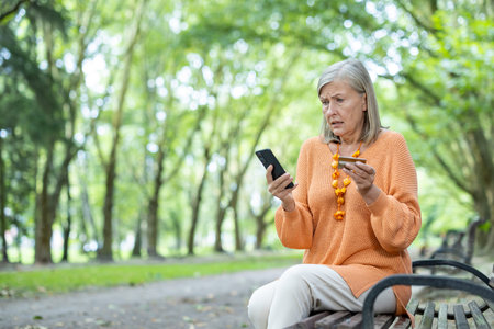 Senior woman outdoors holding smartphone and credit card. She looks concerned and frustrated,...