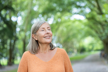 Senior woman standing outdoors in lush green park with eyes closed, radiating peace and contentment. Embracing natures tranquility on a sunny day, embodying relaxation and happiness.の写真素材