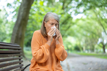 Elderly woman sneezes into tissue while sitting on park. Dressed in cozy sweater, she experiences symptoms of cold or allergies. Natural setting with trees surrounding creates refreshing outdoorの写真素材
