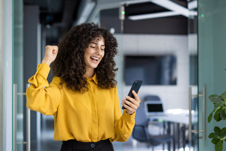 Hispanic businesswoman exudes excitement and triumph checking phone in office. Curly hair, bright yellow blouse, fist pump conveying victory. Modern work setting highlights professional atmosphereの写真素材