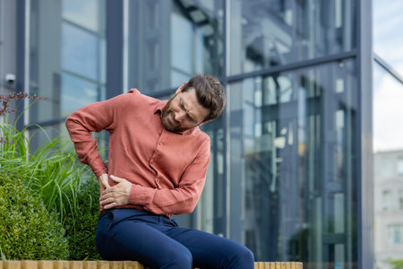 A man in a red shirt is sitting on a bench with his back to the camera. He is holding his stomach and he is in painの写真素材