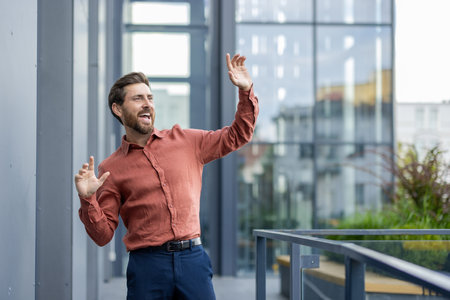 A man in an orange shirt is standing on a balcony. He is smiling and waving. The balcony is surrounded by glass windowsの写真素材