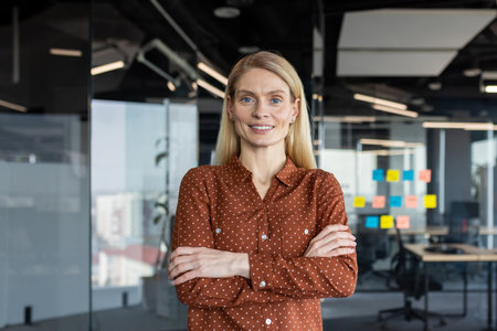 Smiling businesswoman standing with arms crossed in modern office. Professional demeanor exudes confidence and leadership. Ideal representation of success and empowerment in workplace.の写真素材