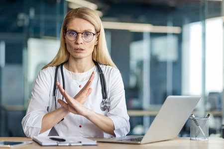 Female doctor in white coat with stethoscope gestures no at laptop desk. She wears glasses, communicates confidently in modern office, expressing authority and professionalism.の写真素材