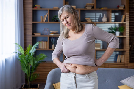 Elderly woman examines stomach with concerned expression. Emphasis on health, wellness, aging, and self-reflection. Scene captures thoughtful awareness of body changes and their emotional impact.の写真素材