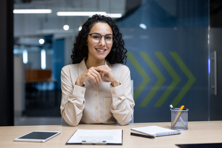 Confident businesswoman wearing glasses smiling at desk with documents, tablet, and stationery in modern office setting, showcasing professionalism and positive work attitude.の写真素材