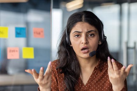 Frustrated customer service representative wearing headset gestures with hands, expressing confusion. Office setting with sticky notes in background, showing challenging communication moment.の写真素材